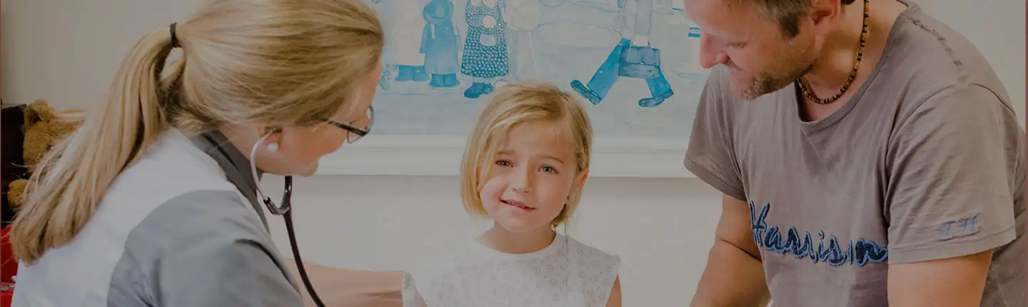 Enfermera examinando a una niña con el fonendoscopio, acompañada de su padre<br/><br/>
