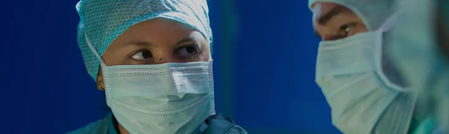 Primer plano de dos especialistas con pijama, gorro quirúrgico y mascarilla.<br/><br/>